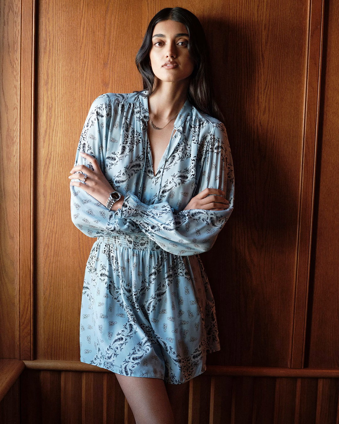 Young woman in a light blue floral long-sleeved short dress standing with arms crossed against a wooden panel wall