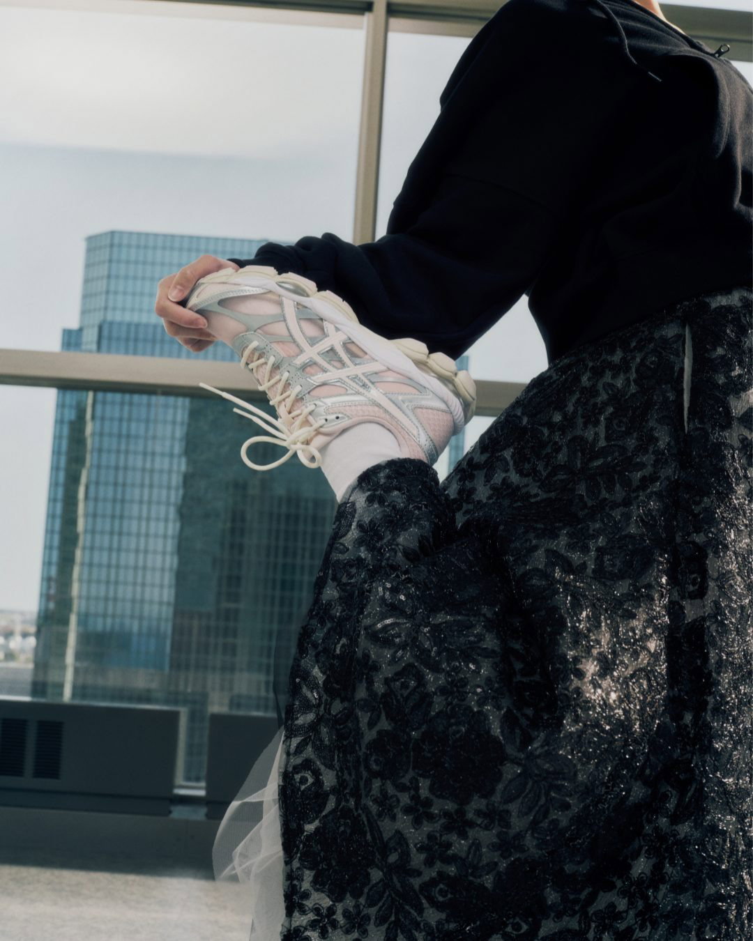 Person in black floral skirt and hoodie holding up white pastel trainers and socks by a floor-to-ceiling city high-rise window