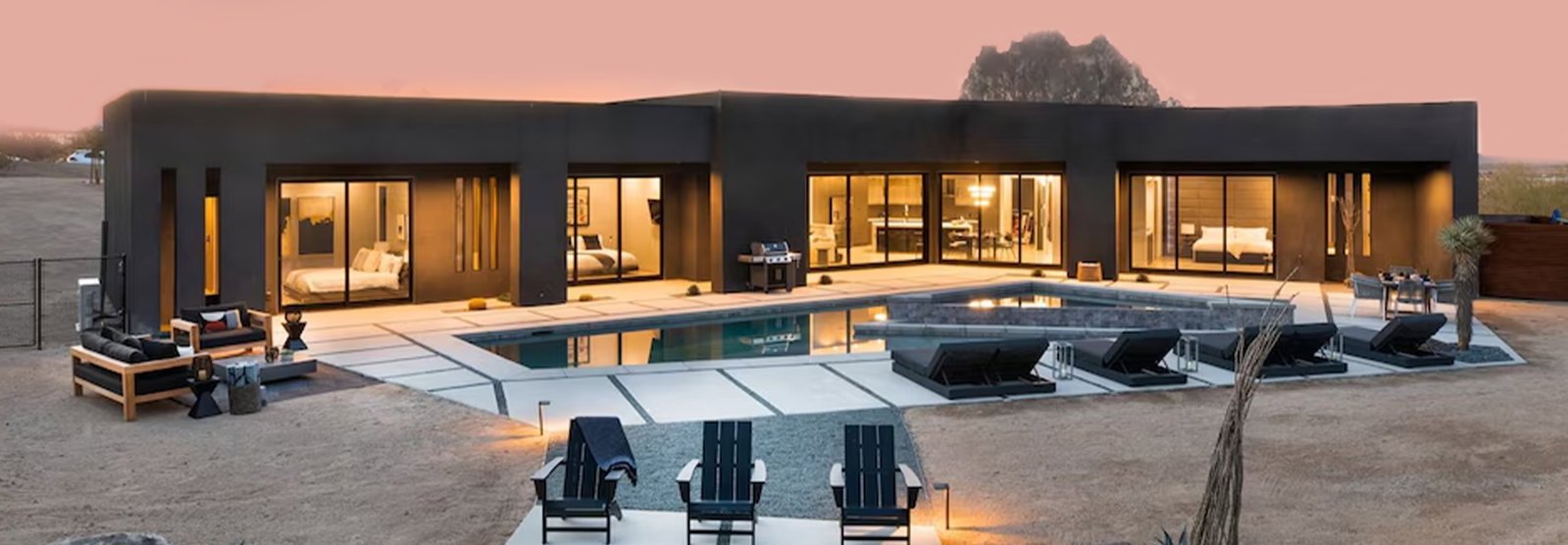 Contemporary desert home with illuminated interiors, expansive pool, and outdoor lounge seating.