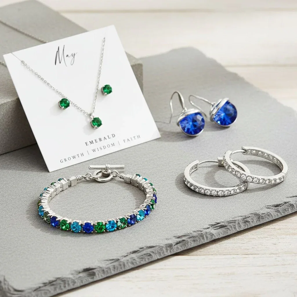 Callura jewelry collection featuring emerald necklace, blue drop earrings, colorful gemstone bracelet, and diamond rings displayed on gray surface.