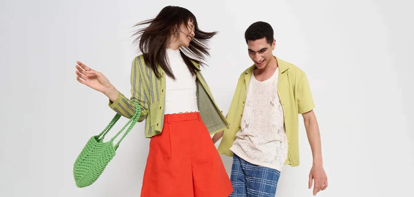 Deux modèles posent dans des tenues d'été colorées : la femme porte un cardigan rayé vert, une jupe rouge et un sac en tissage vert ; l'homme porte un débardeur beige et un short à carreaux bleu.