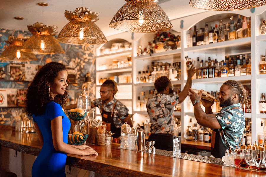 A woman in a vibrant blue dress stands at an elegant, upscale resort bar while bartenders craft cocktails.