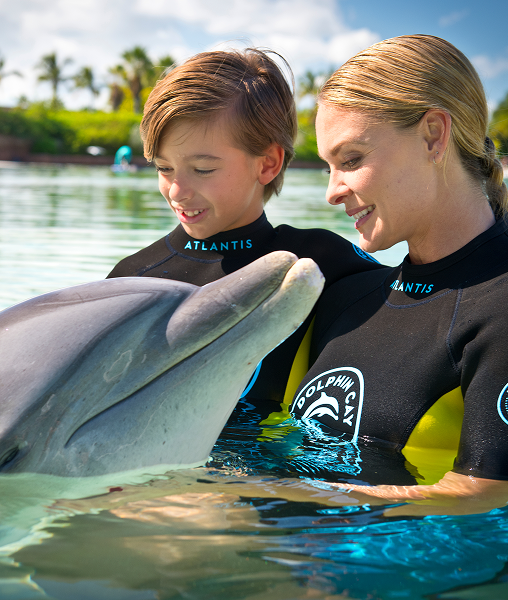 A woman and young boy wearing wetsuits smile together while interacting with a dolphin in a lagoon.