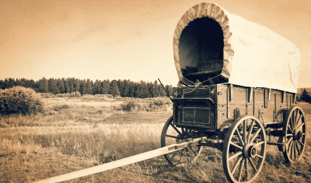 A covered wagon stands in an open, grassy field with a forest in the background. The photo has a sepia tone, giving it a vintage appearance.