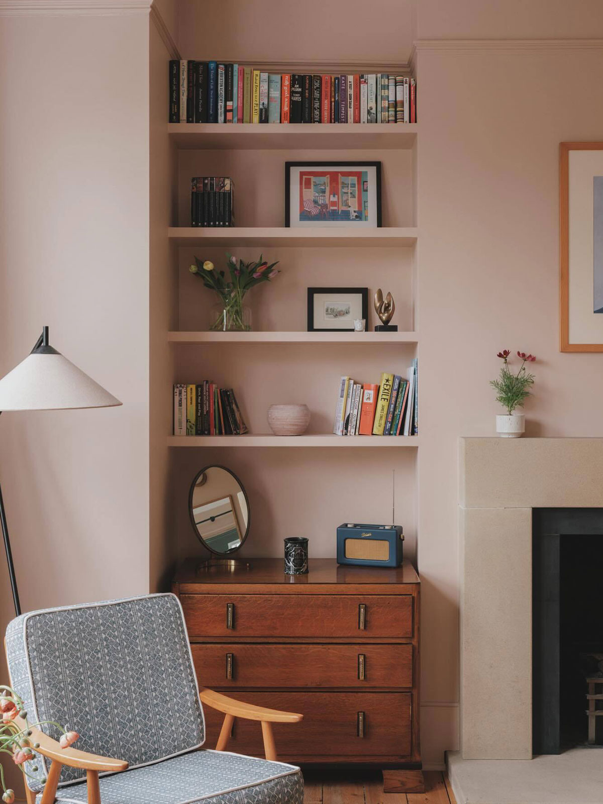 Shelved corner with books, framed art, and a wooden chest of drawers beside a stone fireplace.