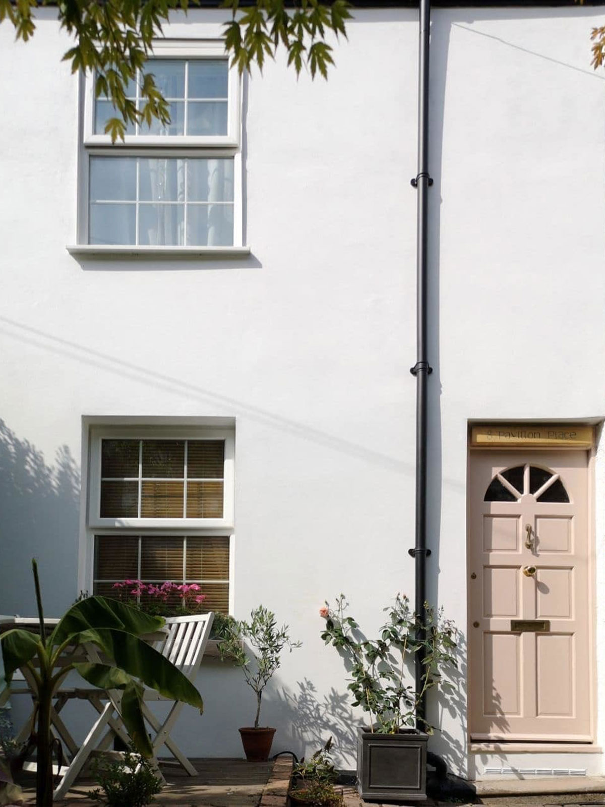 White-painted cottage with a pale pink front door, potted plants, and a small outdoor seating area.