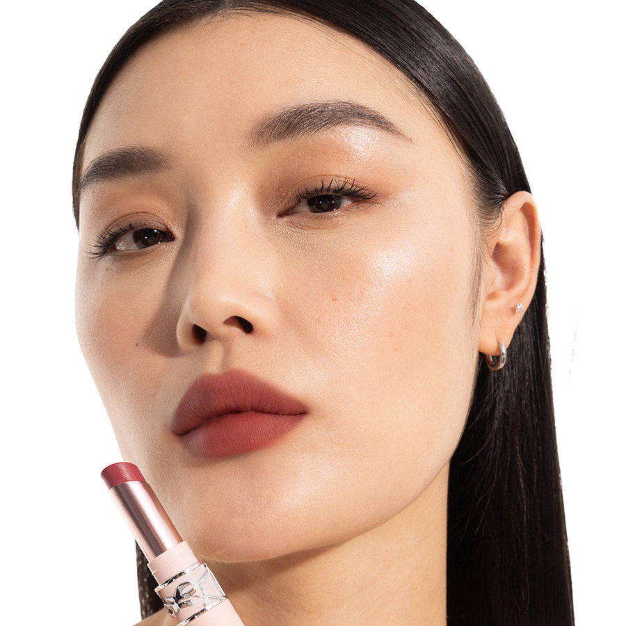 Close-up of an Asian woman showcasing a matte rosewood lipstick shade, holding the matching YSL lipstick.
