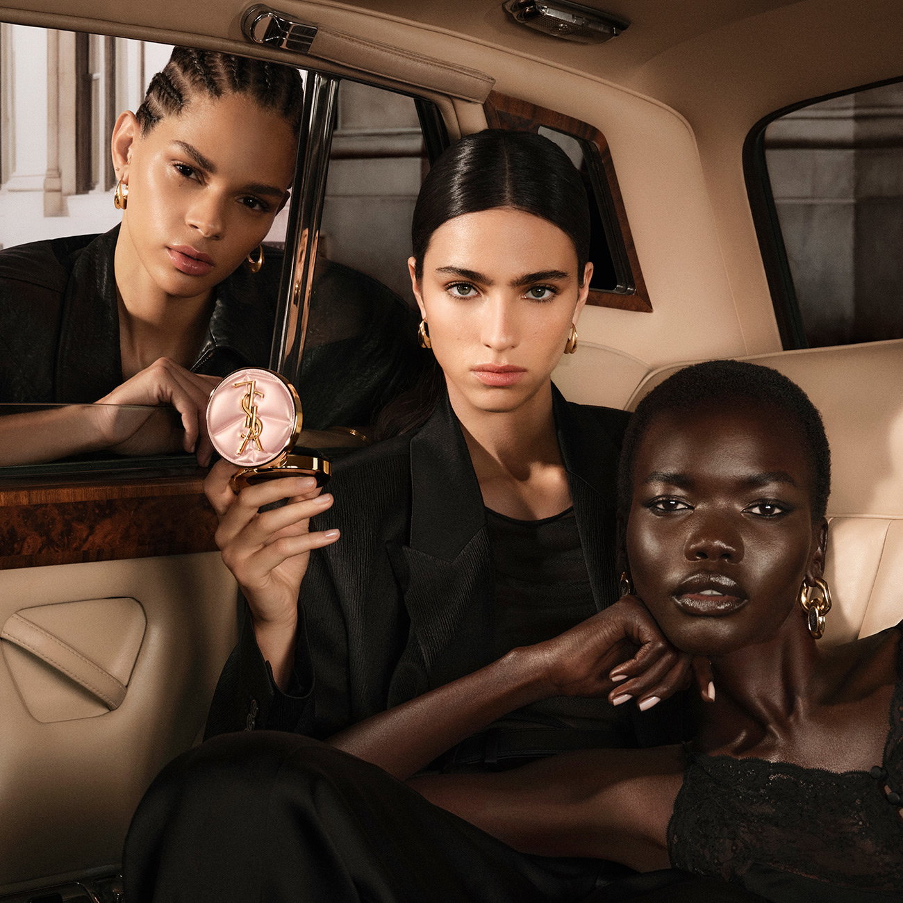 Three elegant women posing inside a luxury vintage car, with one holding a pink quilted YSL cushion compact.