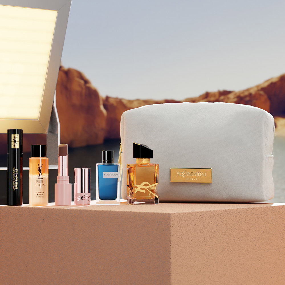 A collection of YSL beauty products, including perfumes, a lipstick, a serum, and a mascara, arranged on a stone block next to a beige makeup bag.