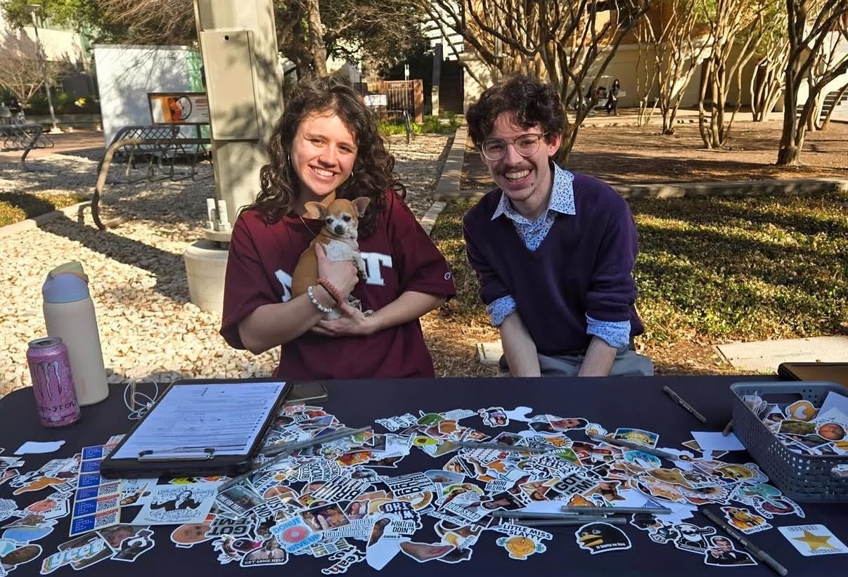 Two people sitting and smiling at a table with lots of stickers spread out on it, a clipboard on the left, an Owala water bottle, and a monster can. Person on the left is holding a chihuahua in their arms.