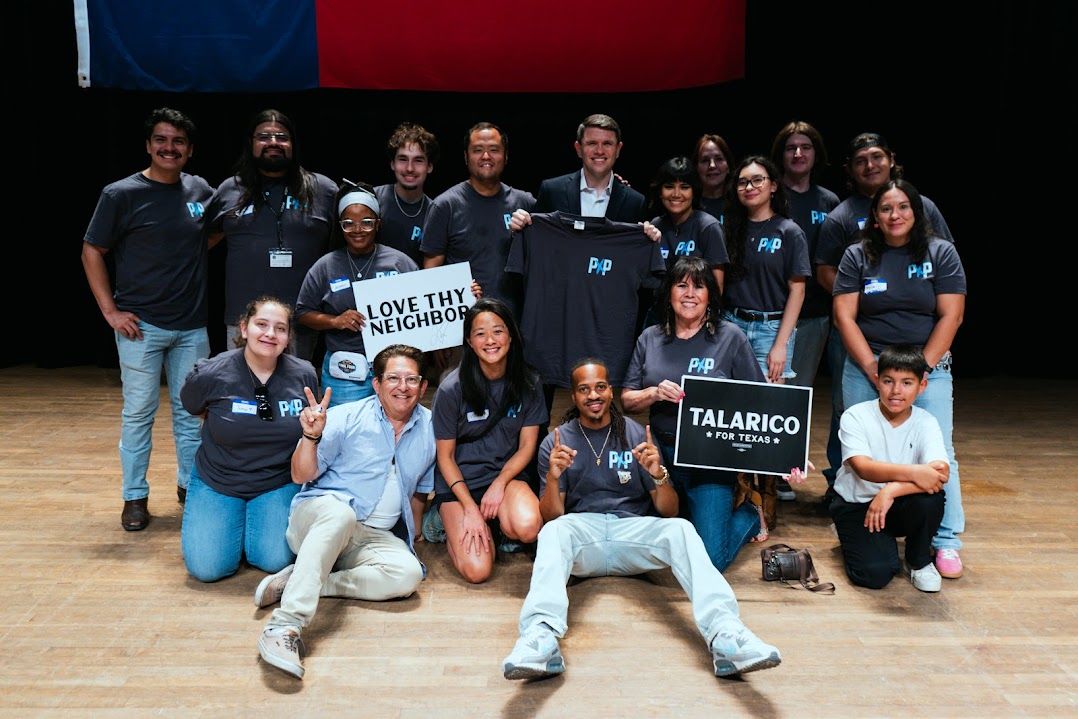 Image of 15 Powered by People volunteers and two more individuals standing with James Talarico on stage. One volunteer, third from the left, is wearing a fanny pack and holding a sign that says "LOVE THY NEIGHBOR." Another volunteer is kneeling to the right of James, holding a sign that says "TALARICO FOR TEXAS"