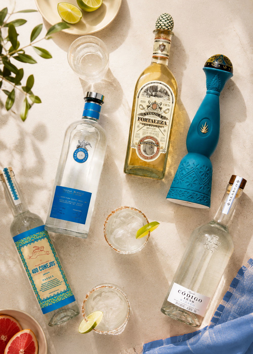 Overhead flatlay of assorted premium tequila bottles, two salt-rimmed lime cocktails, lime wedges, grapefruit and glassware on a light surface