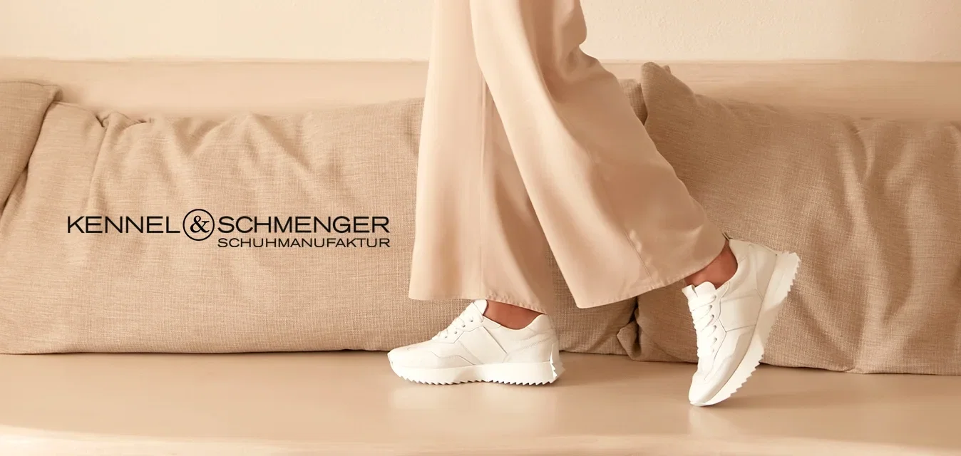 Beige pak met een losvallend colbert en broek met brede pijpen, gecombineerd met witte sneakers. Geplaatst in een minimalistische, neutrale ruimte.