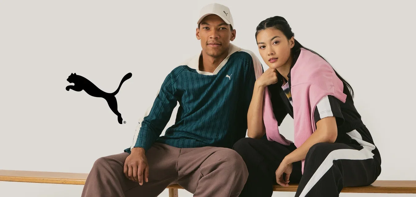 Twee modellen zitten op een houten bank. De man draagt een groen gestreept shirt en een paarse broek, terwijl de vrouw een zwarte jumpsuit met roze accenten heeft.