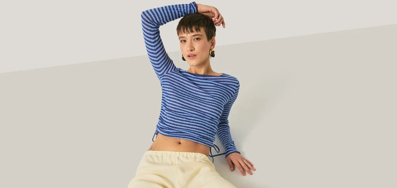 Jonge persoon die achterover ligt met één arm achter het hoofd, gekleed in een blauw gestreept shirt met lange mouwen en lichtbeige broek tegen een effen achtergrond.