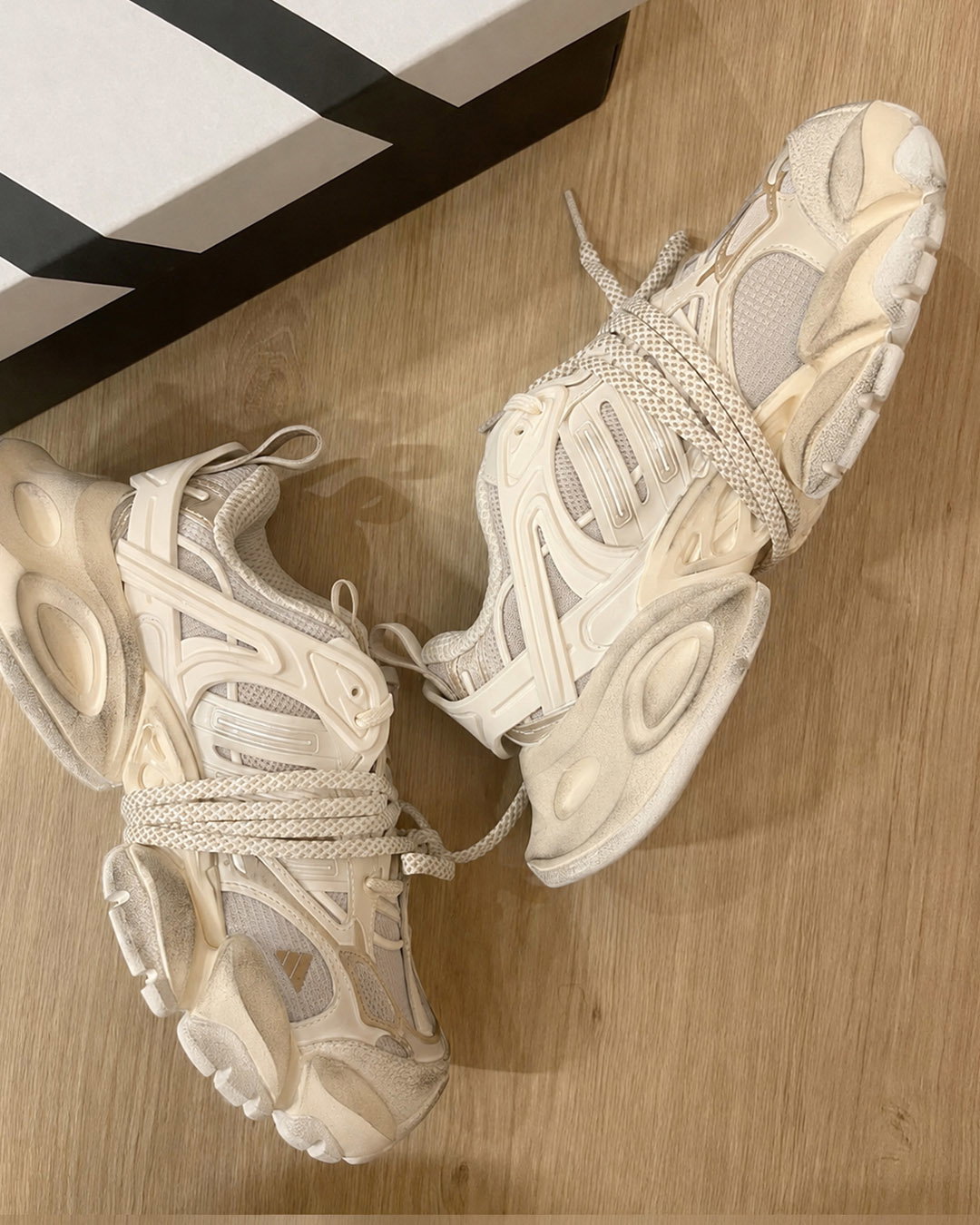 Pair of cream beige chunky athletic trainers with thick textured soles and patterned laces on a light wooden floor beside a black-and-white shoebox