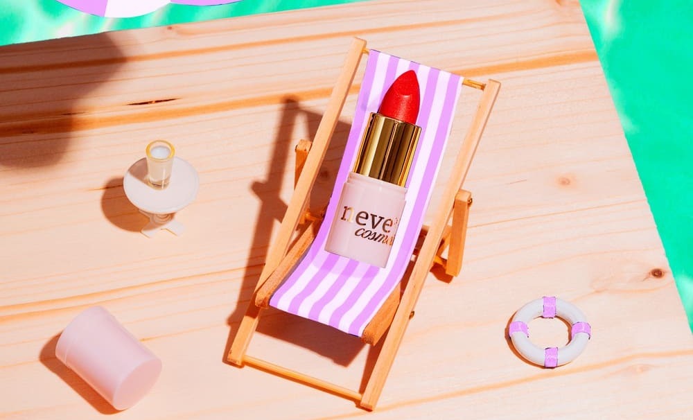 Red lipstick in a gold tube labelled 'never' resting on a pink-and-white striped deckchair on a wooden poolside with a mini lifebuoy and cup