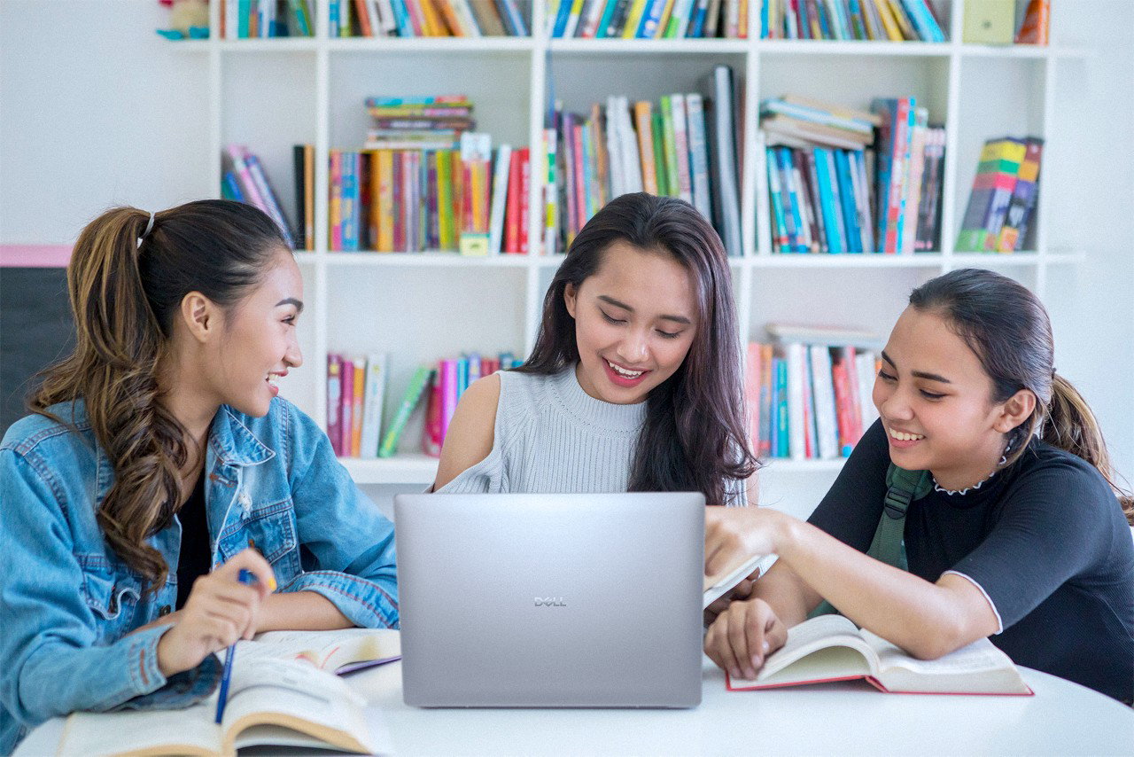 Image of three young women sitting together in a library, studying and looking at a laptop