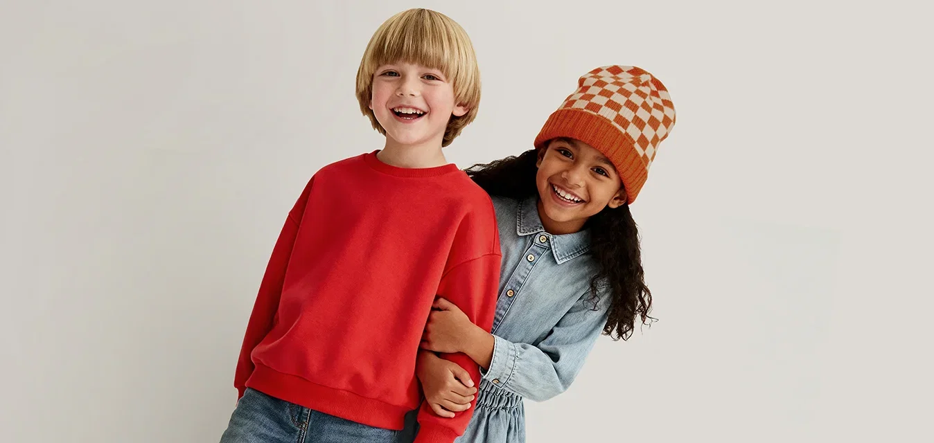 Twee kinderen staan dicht bij elkaar, glimlachend; de ene draagt een rode trui en jeans, de andere een blauwe jurk en een oranje geruit hoedje.