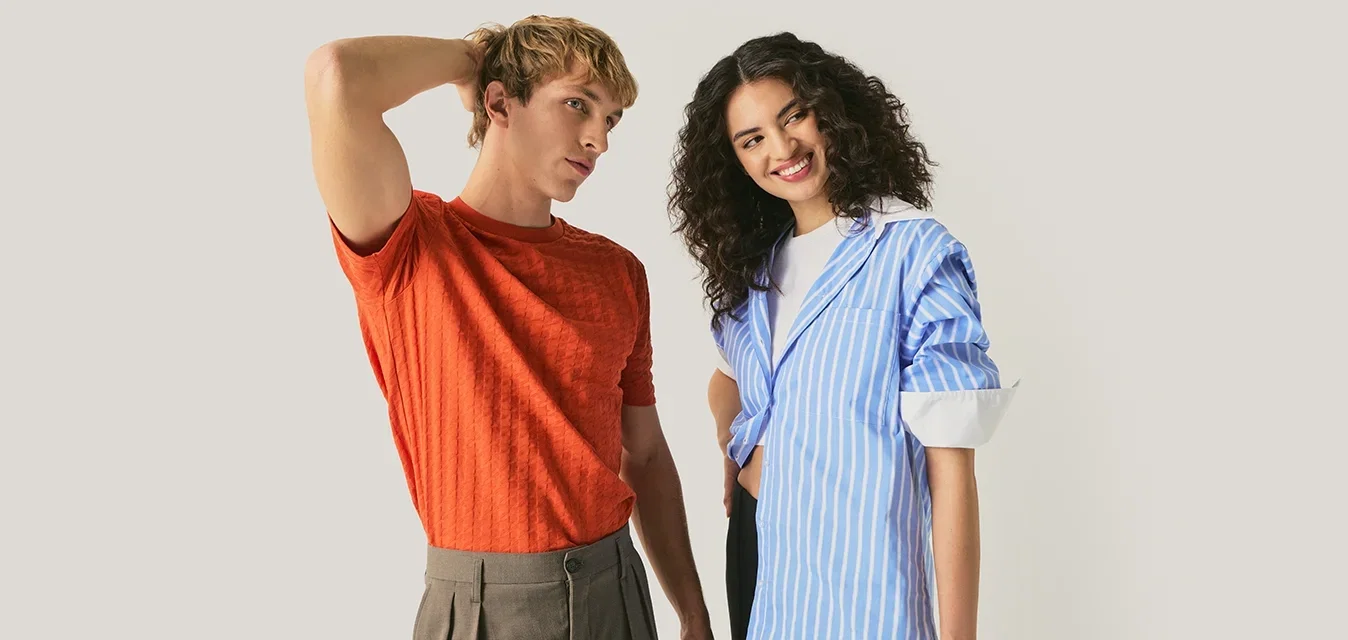Jonge man in oranje shirt en bruine broek staat met hand op hoofd, vrouw in blauw gestreept shirt lacht met hand op heup, neutrale achtergrond.