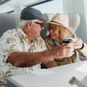 An older couple sits together on a modern train, smiling warmly at each other while holding takeaway coffee cups. The man wears a cap and patterned shirt, and the woman wears a straw hat and glasses. Bright natural light fills the cabin, creating a relaxed and cheerful travel atmosphere. The scene conveys companionship, leisure, and comfortable transportation.