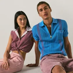 Un uomo con una camicia blu a maniche corte e pantaloncini rosa è seduto accanto a una donna con una maglia rossa a righe e pantaloni rosa su uno sfondo chiaro e uniforme.