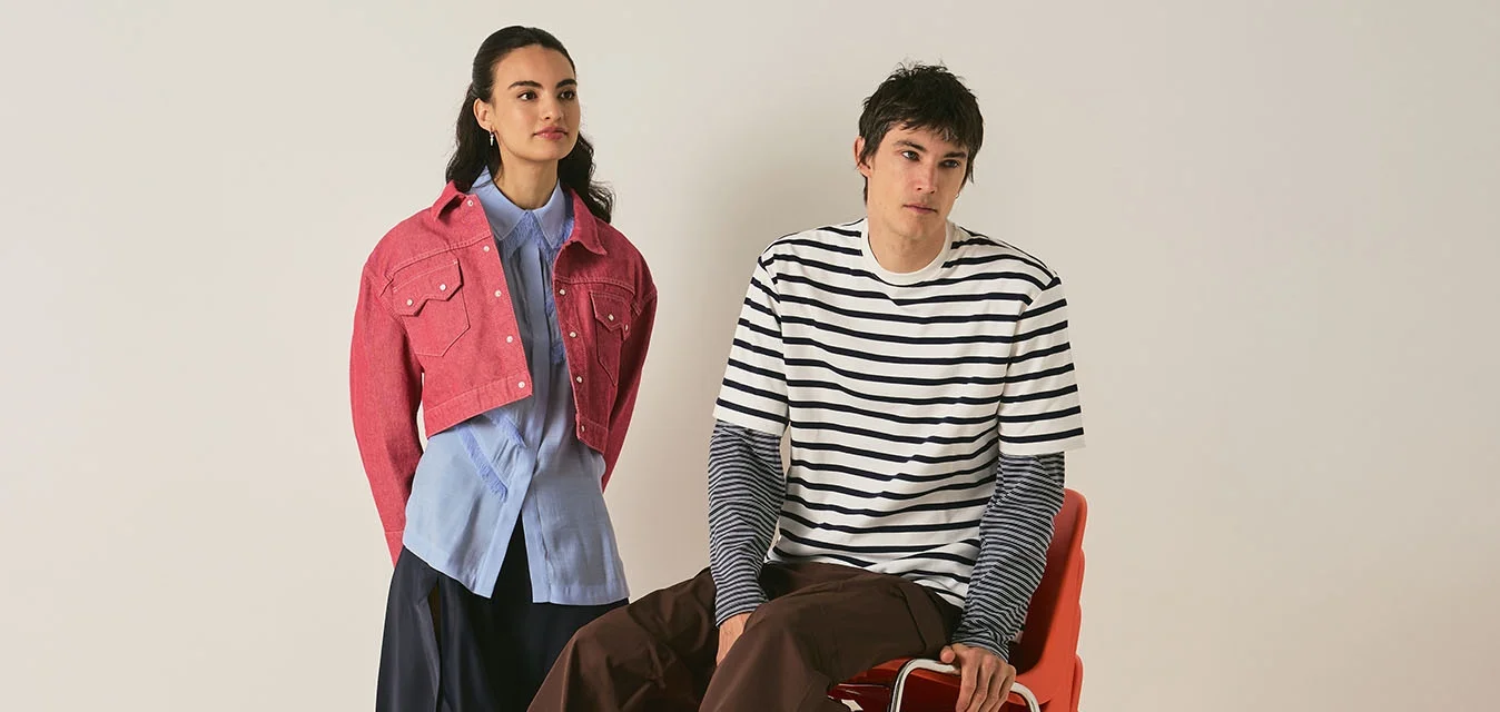 Donna in piedi con giacca rosa, camicia blu e gonna nera; uomo seduto su sedie arancioni impilate, indossa una camicia a righe e pantaloni marroni su uno sfondo semplice.