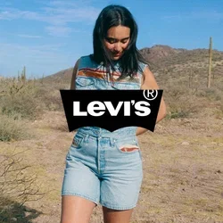 Donna con tuta corta in denim senza maniche in piedi in un deserto con cactus e montagne sotto un cielo azzurro limpido.