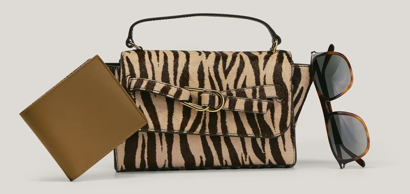 Portafoglio in pelle marrone, borsa zebrate con fibbia e occhiali da sole in tartaruga disposti su sfondo neutro.