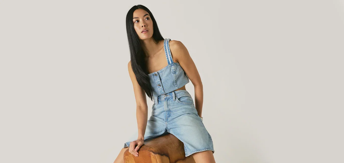 Donna con abito di jeans senza maniche seduta di lato su un cavallo con maniglie da palestra in legno, guarda verso sinistra, un piede in una scarpa nera sul pavimento.
