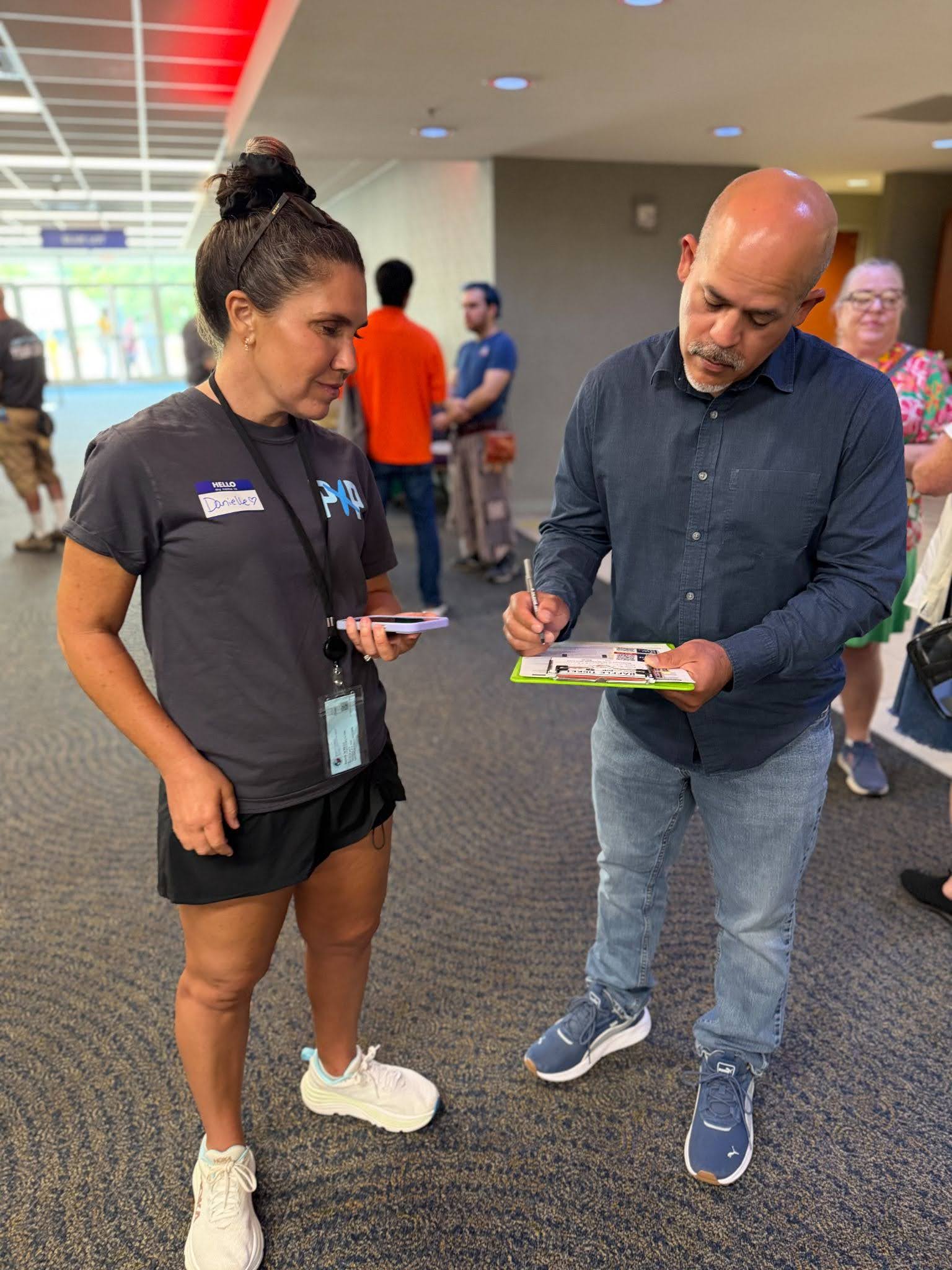 A Powered by People volunteer wearing a gray PXP shirt and lanyard (left) helping someone writing on a clipboard (right) get registered to vote.
