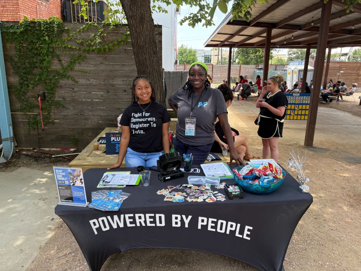 Two Powered by People volunteers standing at a table with a black table cover that says "POWERED BY PEOPLE" on it. On the table there are stickers, a bowl of snacks, brochures, pens, a fan, and clipboards with voter registration papers attached to them. 