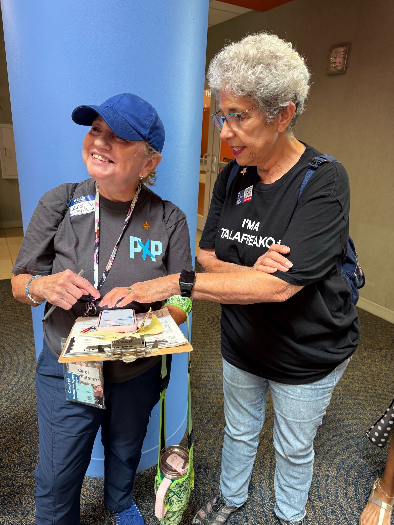 A Powered by People volunteer wearing a gray PxP tee shirt holding a clipboard with their phone resting on top of it (left), smiling towards something off camera; another person in the photo is wearing a black tee shirt that says "I'M A TALAFREAKO" (right)