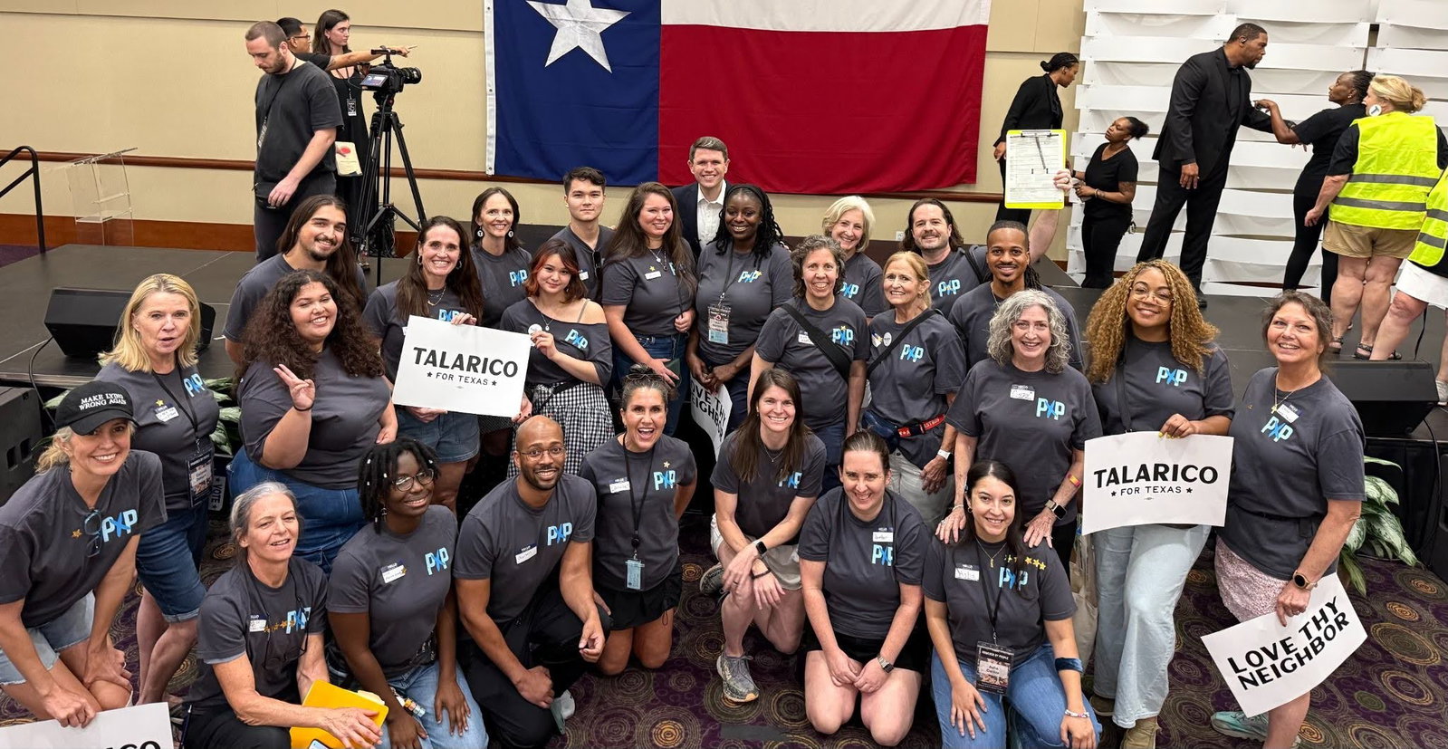 Group of Powered by People volunteers standing in front of a stage with the Texas flag in the background. James Talarico stands in the center of group in the back row. Two volunteers are holding signs that say "TALARICO FOR CONGRESS" and one volunteer is holding a sign that says "LOVE THY NEIGHBOR." 