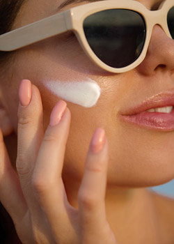 Close-up of woman in cream sunglasses applying sunscreen to cheek with pink manicured nails, summer skincare