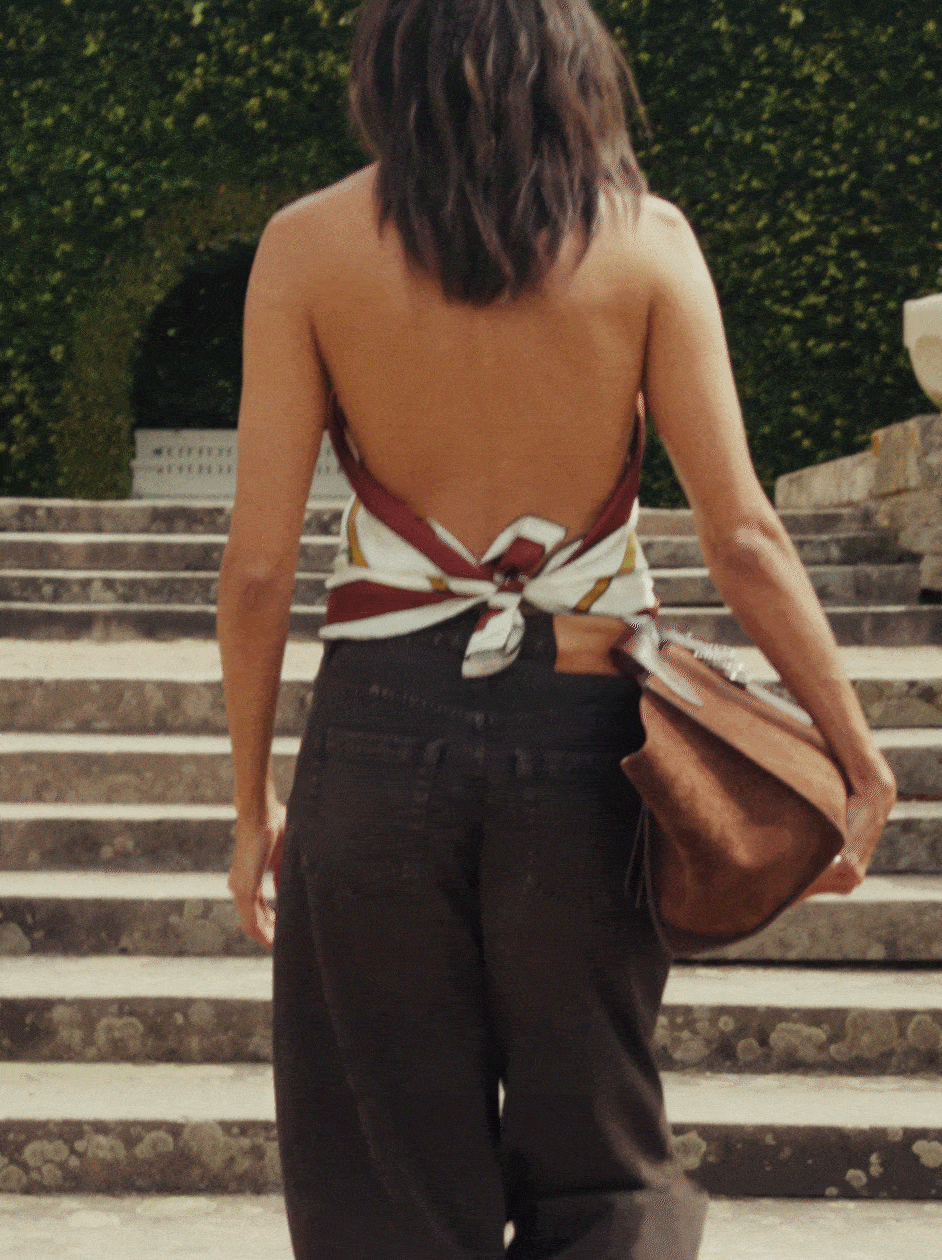Frau von hinten mit offenem Rücken, geht in weiter Jeanshose eine Steintreppe hinauf, trägt ein Seidentuch in Bordeaux und Weiß als Top gebunden sowie eine große Ledertasche in Cognac, im Hintergrund eine grüne Hecke.