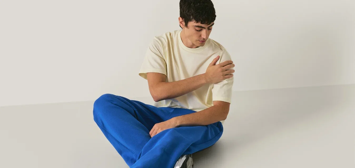 Joven sentado en el suelo con una camiseta beige y pantalones azules, sujetándose el hombro izquierdo y mirando hacia abajo en una habitación minimalista.