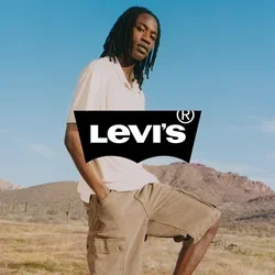 Hombre con rastas que lleva pantalones cortos beige y camisa blanca, equilibra un pie sobre una silla plegable amarilla en un paisaje seco y montañoso bajo un cielo azul.