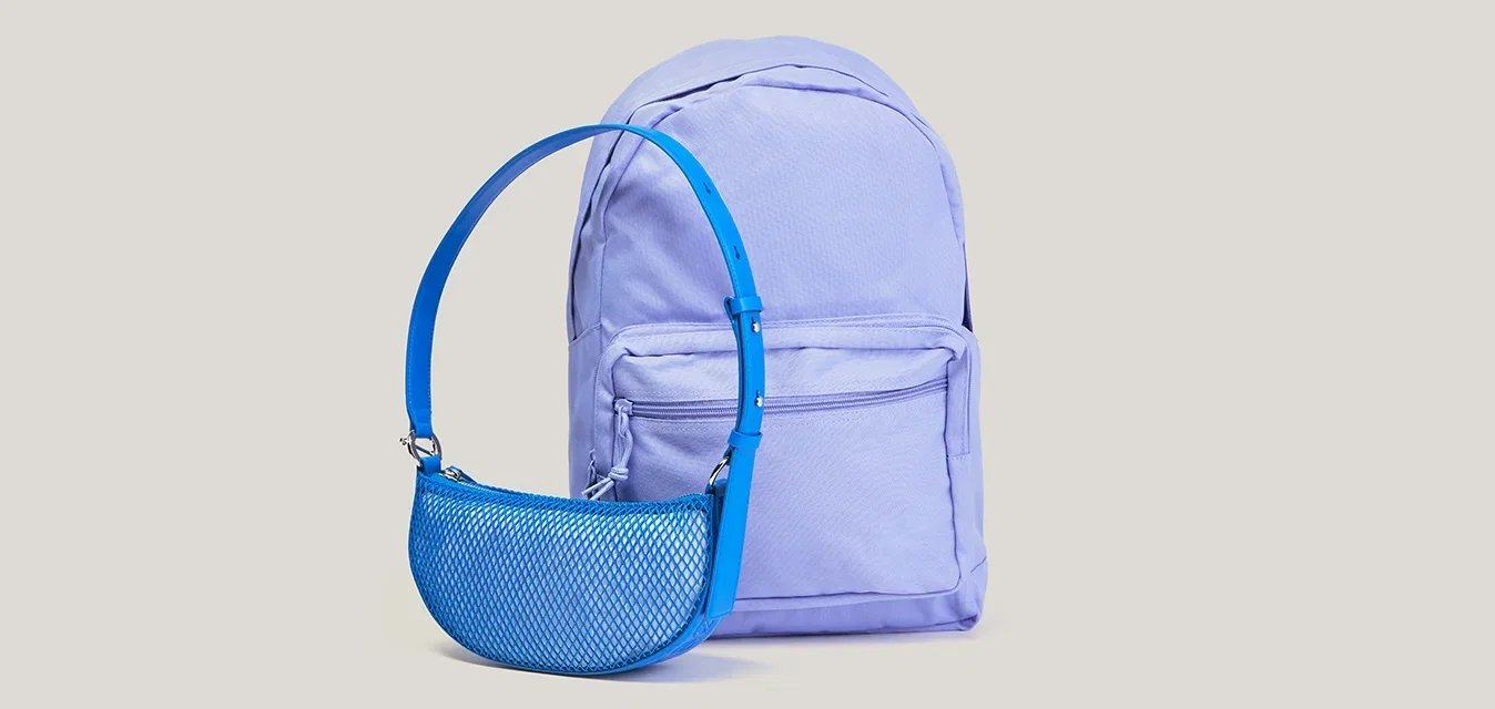 Mochila de color púrpura claro con bolsillo delantero y pequeña bolsa de malla azul con correa ajustable colocadas sobre un fondo liso.