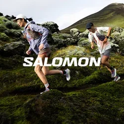 Dos corredores de trail con ropa deportiva navegan por una ladera rocosa y cubierta de musgo con una montaña al fondo.