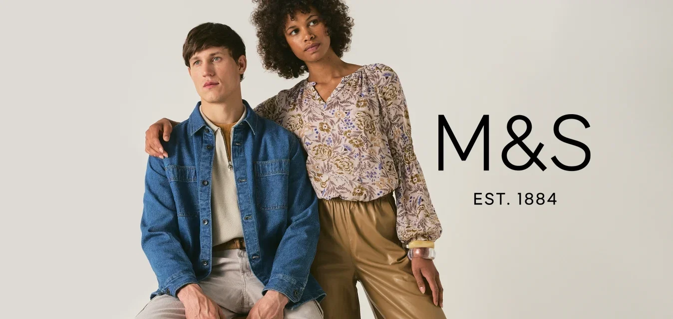 Homme assis sur un tabouret en bois, portant une veste en jean et un pantalon clair ; femme debout, le bras sur son épaule, en blouse fleurie et pantalon ample beige.