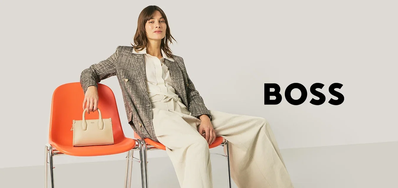 Femme en tenue professionnelle assise de manière détendue sur deux fauteuils orange, tenant un sac à main beige sur un fauteuil, sur un fond clair et uni.