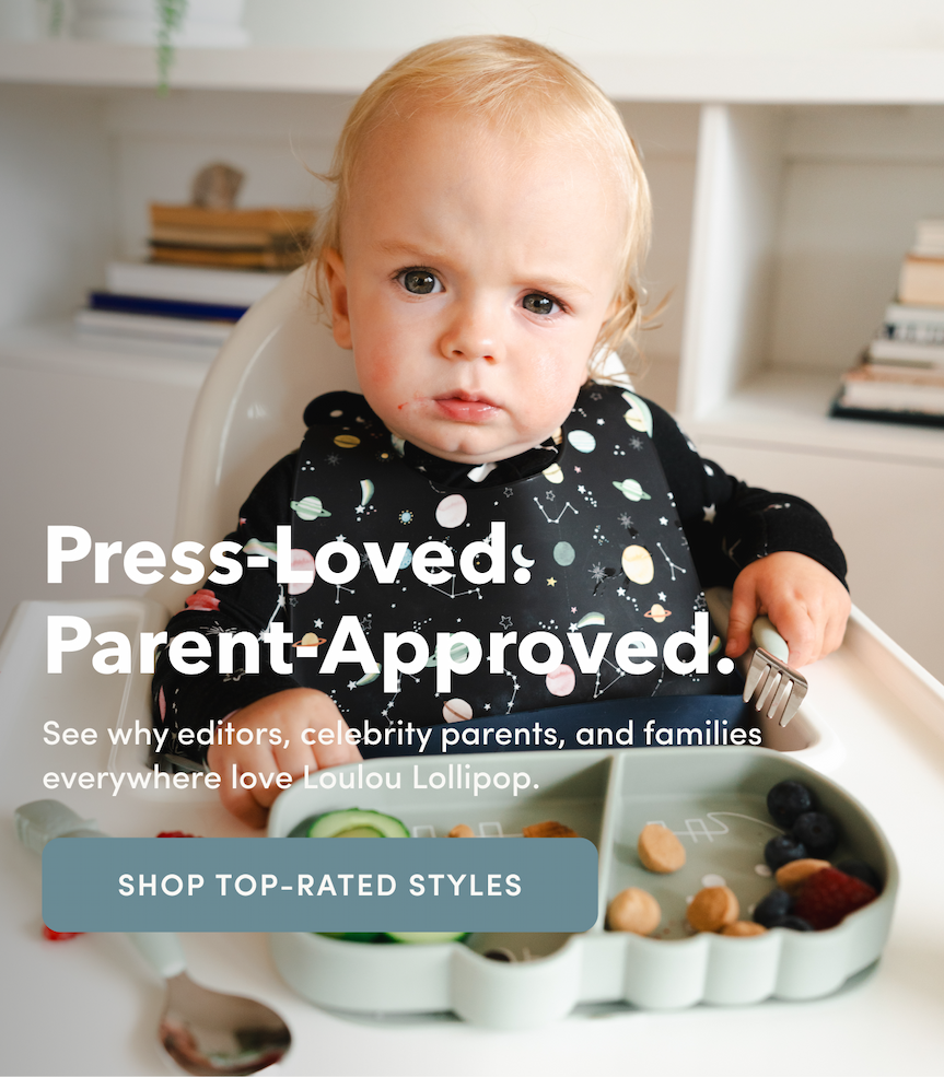 baby sitting in a high chair wearing a black bib and eating off a green alligator shaped silicone plate