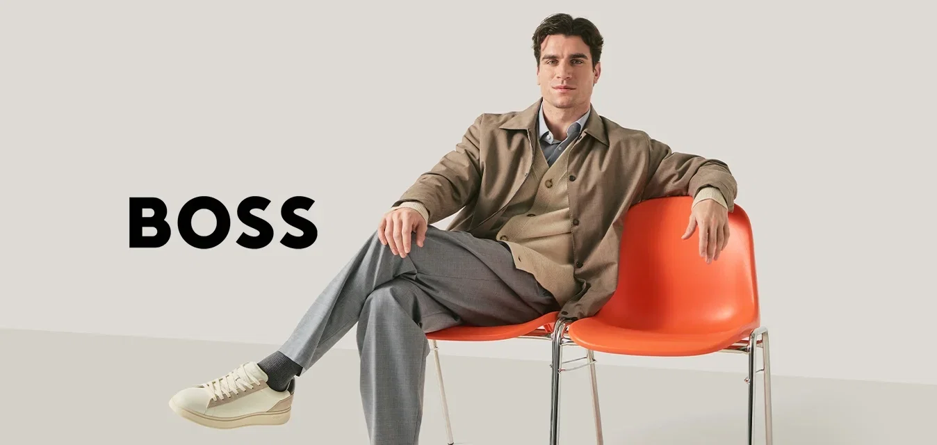 Man in beige blazer en grijze broek zit met gekruiste benen op een van de twee verbonden oranje stoelen tegen een effen achtergrond.