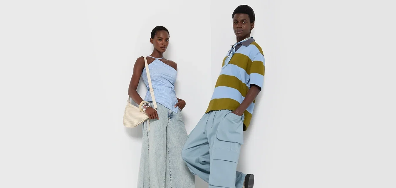 Twee modellen staan tegen een witte muur. De vrouw draagt een lichtblauwe ribbeltrui en brede denim broek. De man heeft een groenblauw gestreept polo-shirt aan met lichtblauwe cargobroek.