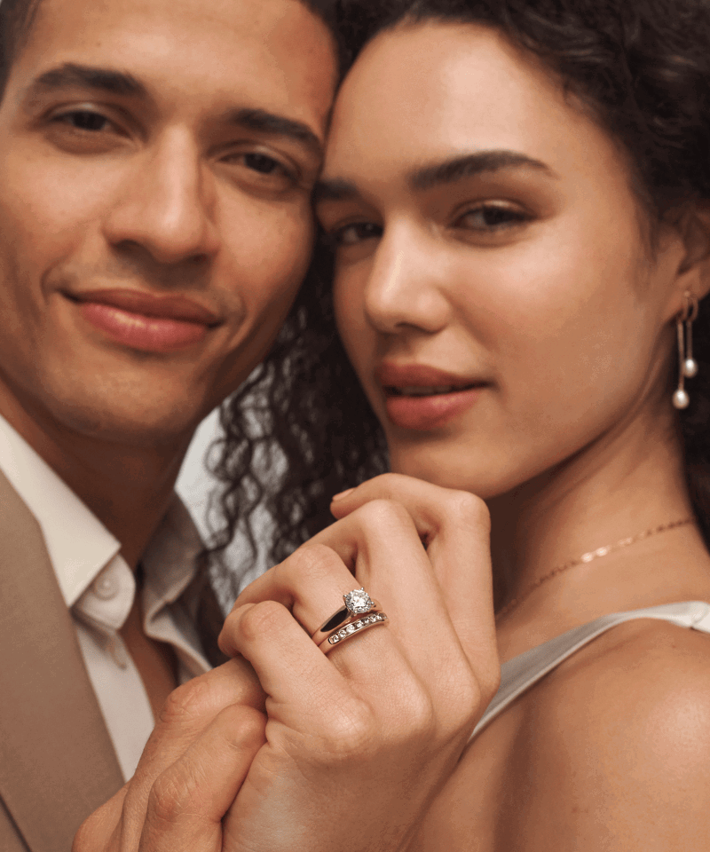Couple holding hands. Woman is wearing diamond bridal rings, pearl drop earrings and yellow gold chain.