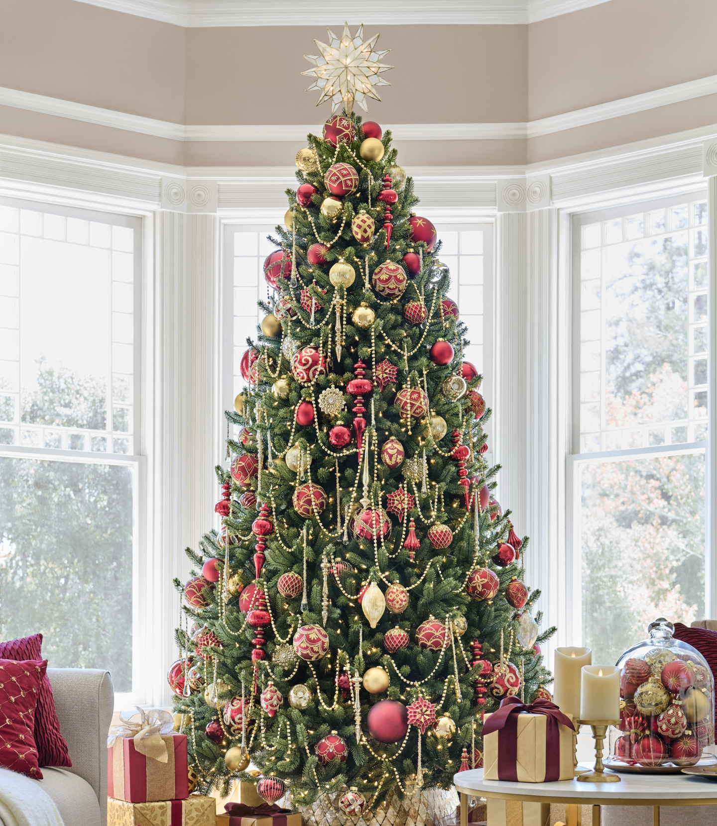 Pre-lit Christmas wreath and garland with pinecones, green foliage, and warm lights displayed on a white mantel with candles and wrapped gifts.