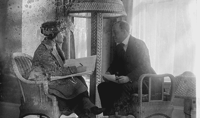 A man and a woman are seated in wicker chairs, engaged in conversation, with a tall lamp between them. The setting appears to be indoors with light coming in through the windows behind them.