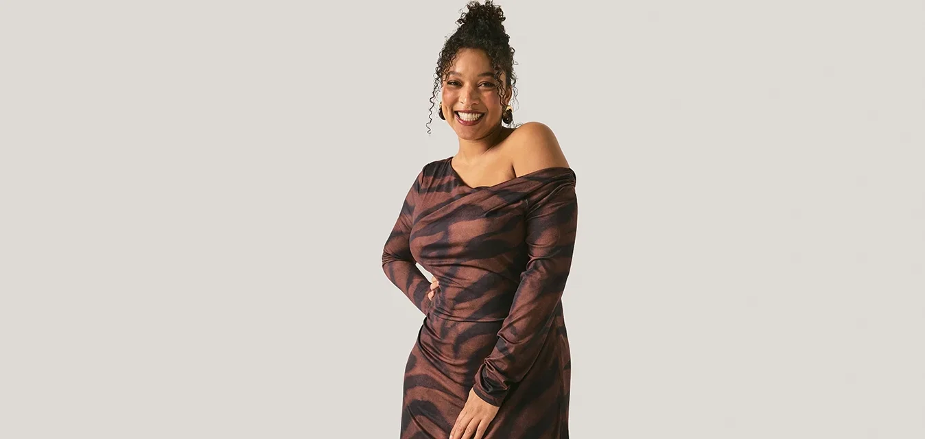 Femme souriante aux cheveux bouclés portant une robe à motifs marron et noir, décolleté sur les épaules, se tenant devant un fond clair uni.