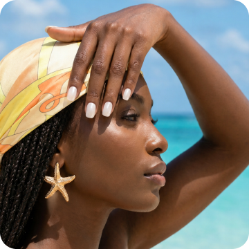 Profile shot of a woman at the beach wearing a patterned headscarf and starfish earrings, touching her forehead with a hand featuring Dashing Diva Pure Porcelain Glaze Effect.